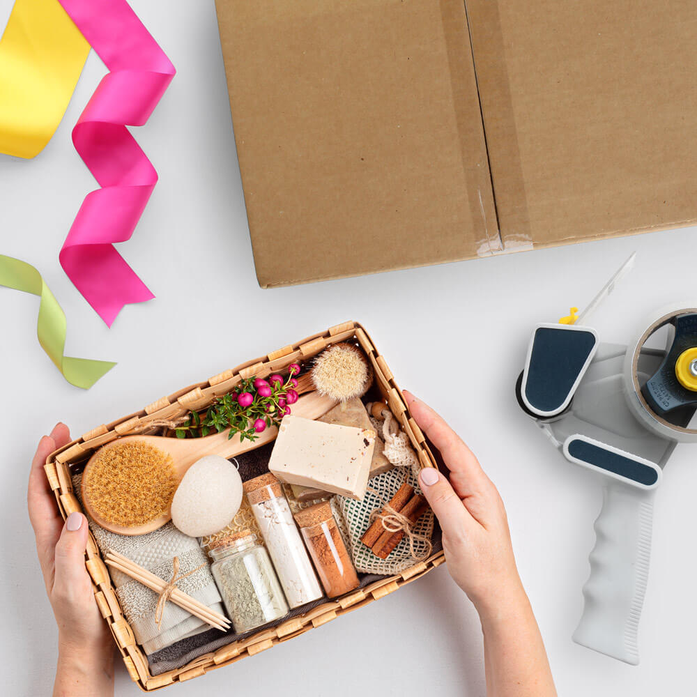 overhead image of hands holding a gift basket alongside a cardboard box, tape dispenser, and ribbon